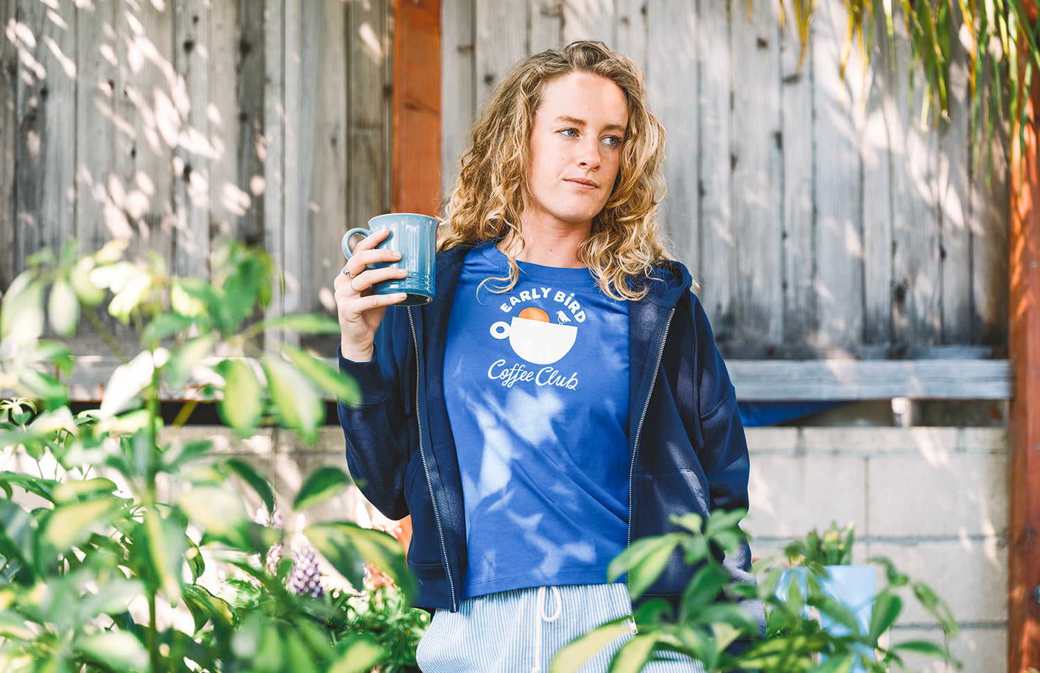 Woman drinking a coffee in a garden with a blue early bird t shirt coffee club